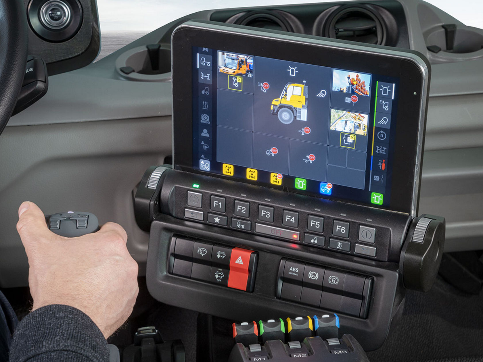 A close-up of a dashboard with a screen showing multiple camera perspectives. One hand holds an operating lever next to the screen.