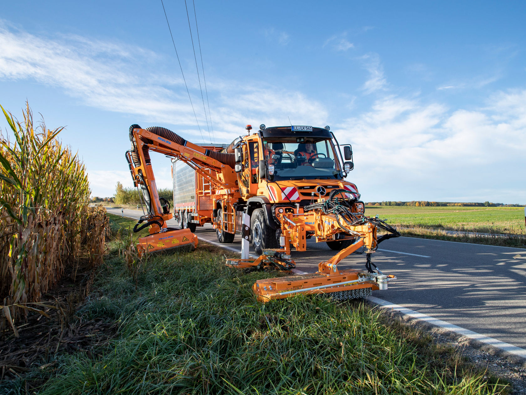 An orange Unimog implement carrier with mower unit mowing grass at the roadside. The vehicle is driving on a road that runs along a field.