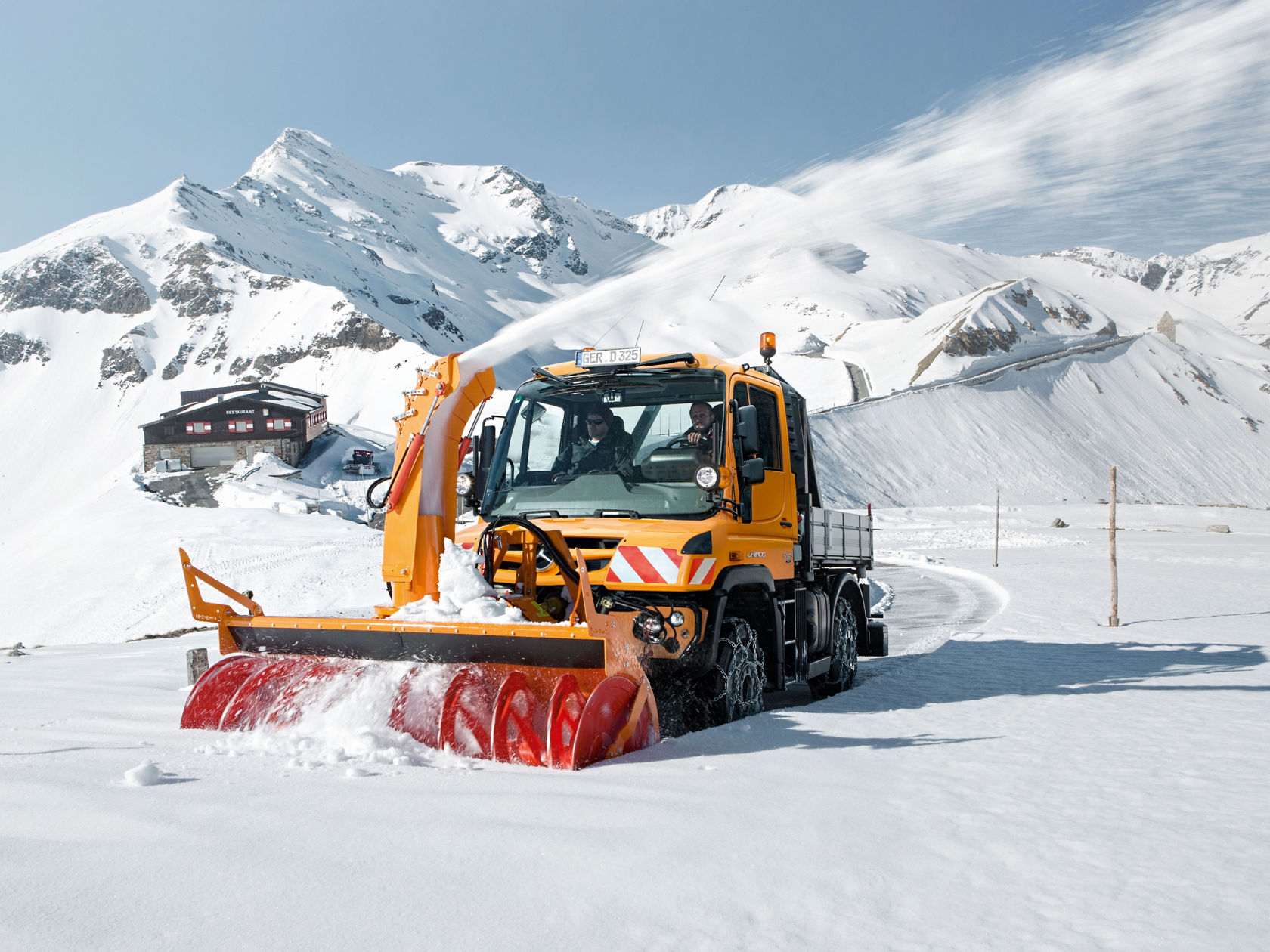 An orange Unimog implement carrier clears a road of snow in a snow-covered mountainous area.