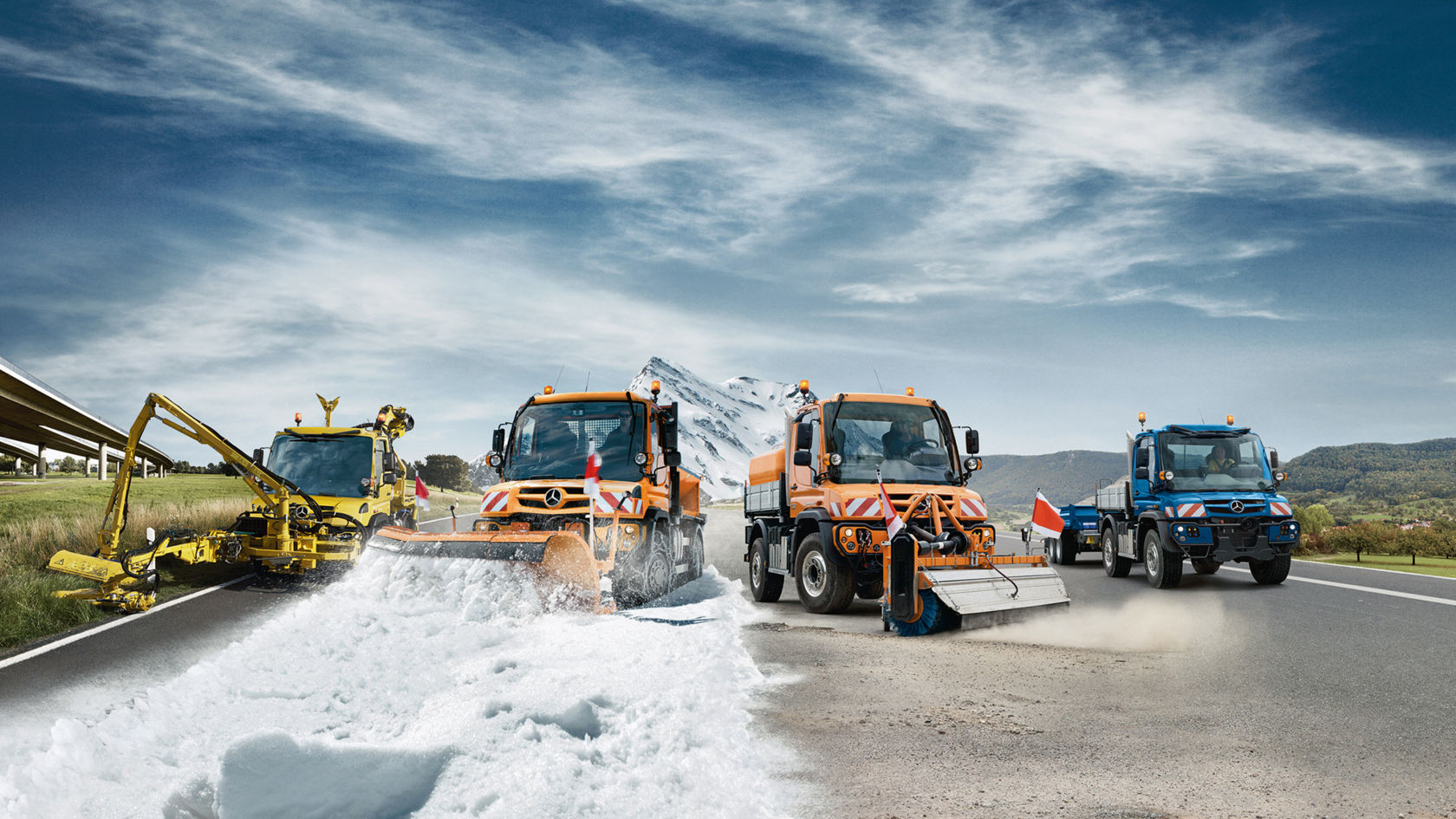 Four Unimog implement carriers in different colours and with different applications stand next to each other on a wide paved road: winter service, road cleaning, agricultural and transport operations. Green meadows and mountains, a snow-covered mountain and a motorway bridge can be seen in the background. 