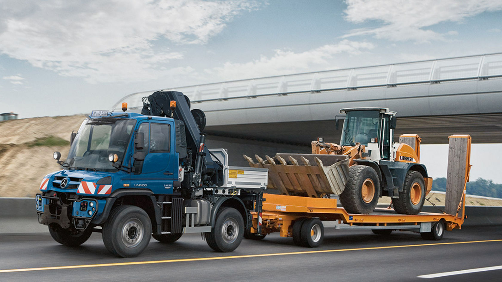 A blue Unimog implement carrier drives under a motorway bridge. It is transporting an excavator.