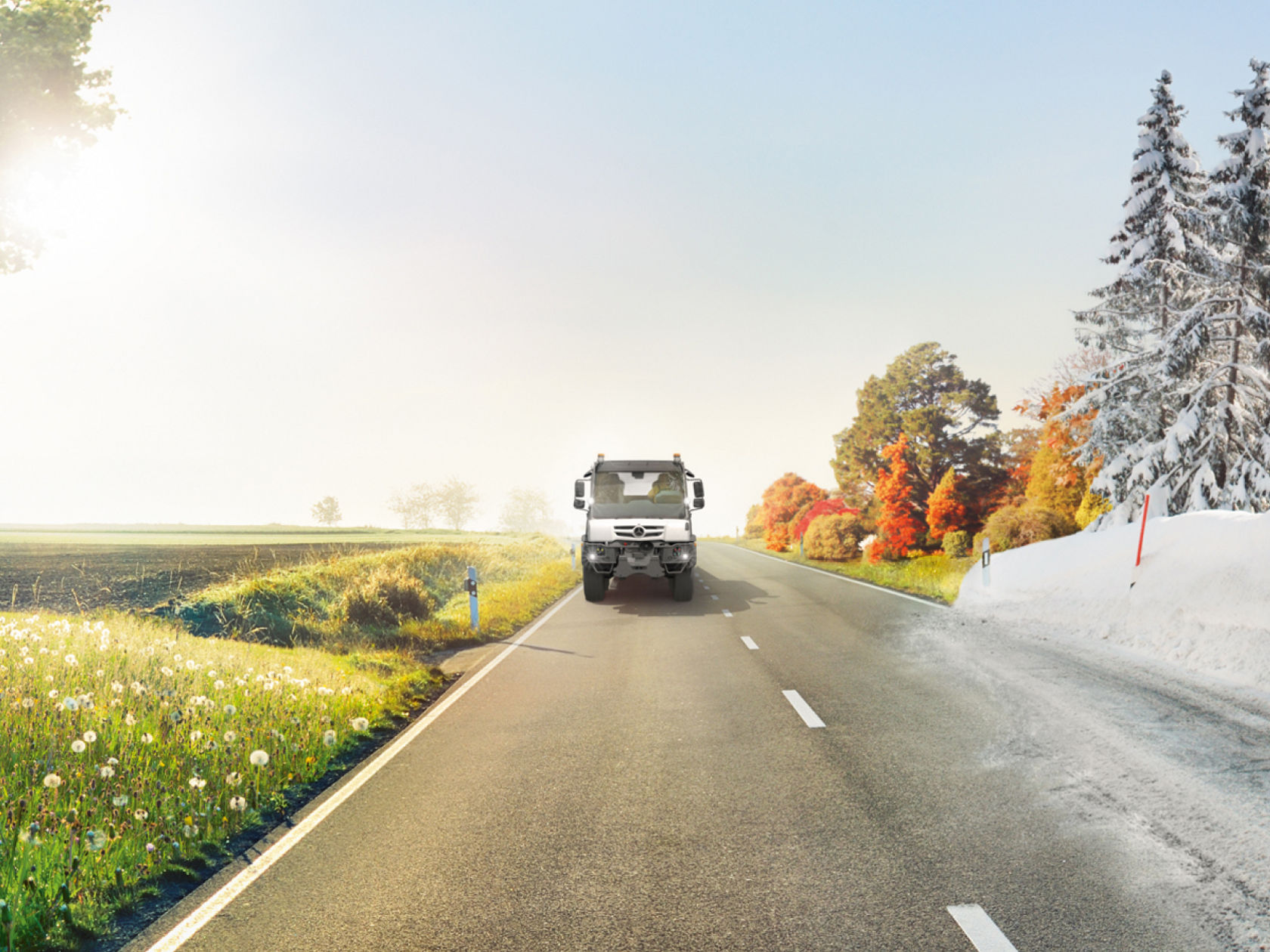 A Unimog drives on a country road that runs through a divided landscape. On the left, a spring meadow blooms, while on the right, winter conditions with snow and snowy trees prevail.