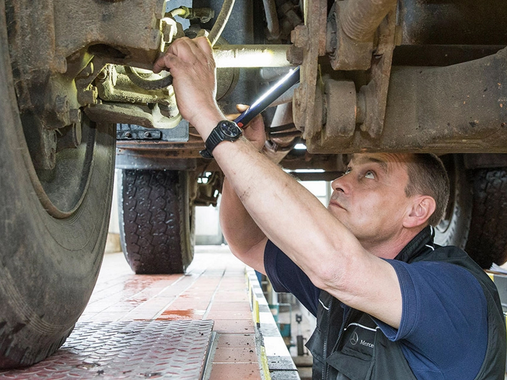 A Mercedes-Benz Trucks mechanic in work clothes checks details on the underbody of a Unimog from below with a lamp. The truck is on a vehicle lift.